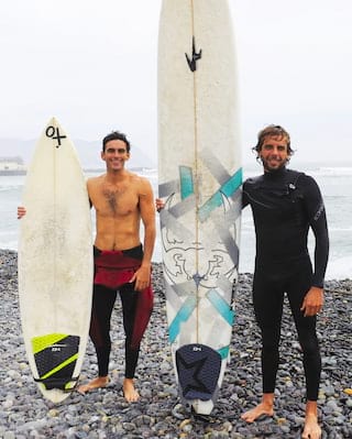 Two men holding surf boards on a stoney beach in Lima