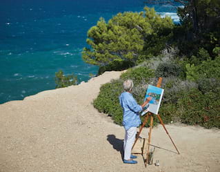 Man painting a canvas on an easel on the beach beside a blue ocean